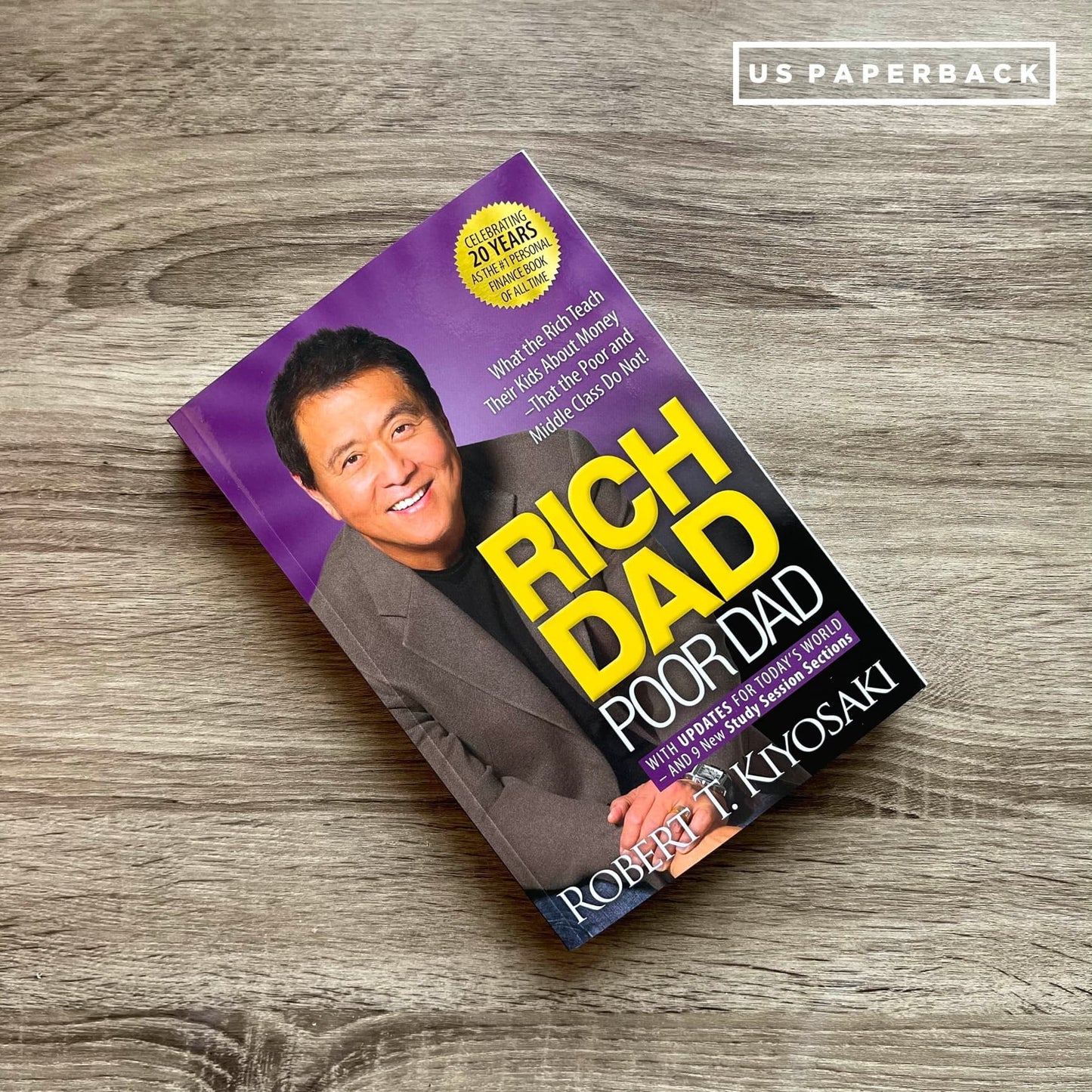 Book cover of "Rich Dad Poor Dad" by Robert T. Kiyosaki. The cover has a purple background with a large yellow title "RICH DAD" and a smaller white subtitle "POOR DAD." A smiling photo of the author, Robert T. Kiyosaki, is featured prominently. A circular badge at the top left reads "CELEBRATING 20 YEARS AS THE PERSONAL FINANCE CLASSIC." The bottom of the cover includes the author's name in white text.

